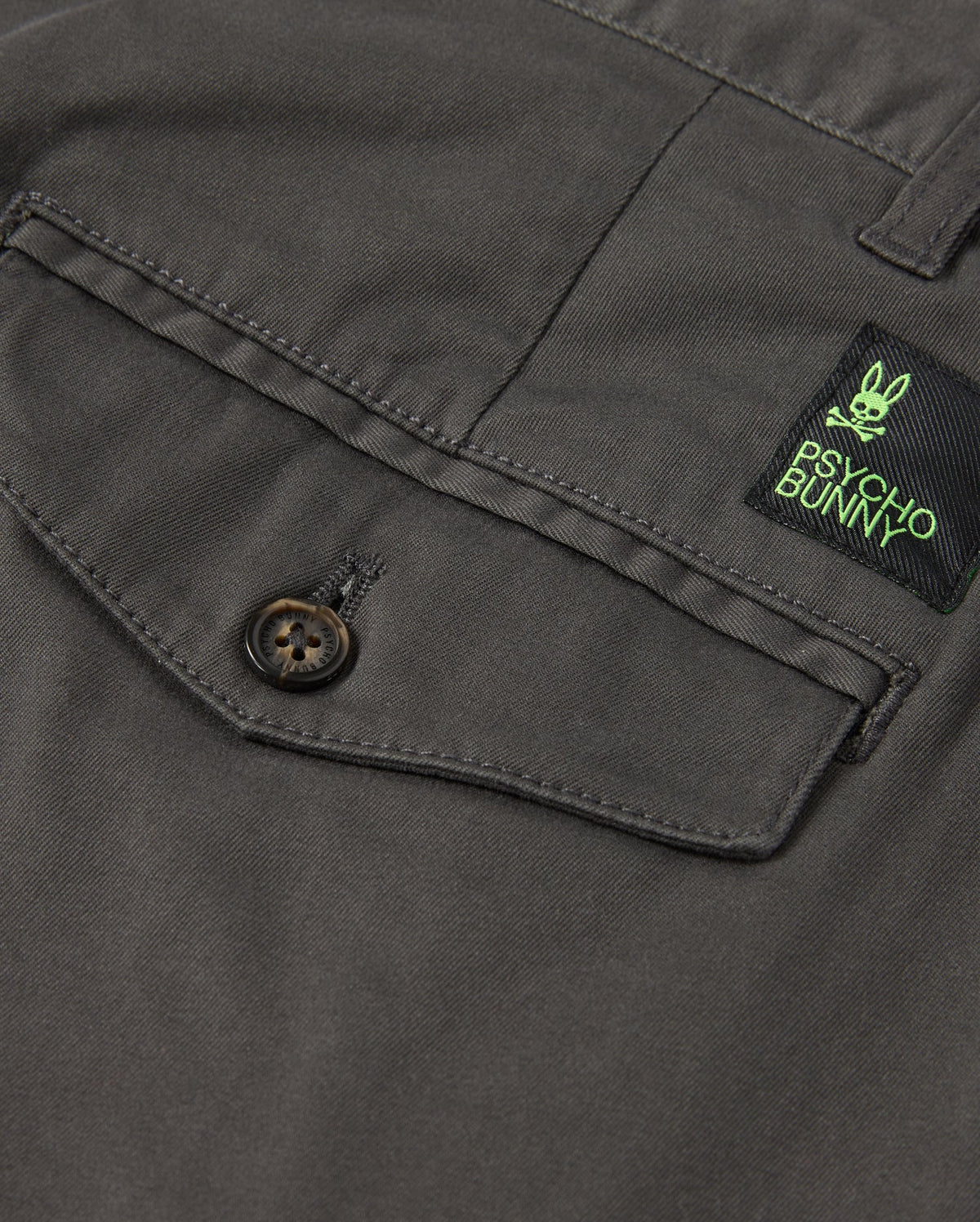 Detailed view of the gray fabric featuring a buttoned pocket, designed for stretch and comfort. Positioned above the pocket is a green and black label showcasing a bunny logo along with "Psycho Bunny," evoking the classic style of the MENS GILMAN CHINO PANT - B6P393Z1WB.