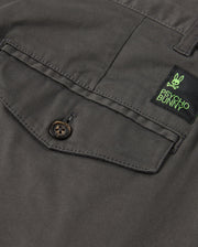 Detailed view of the gray fabric featuring a buttoned pocket, designed for stretch and comfort. Positioned above the pocket is a green and black label showcasing a bunny logo along with "Psycho Bunny," evoking the classic style of the MENS GILMAN CHINO PANT - B6P393Z1WB.