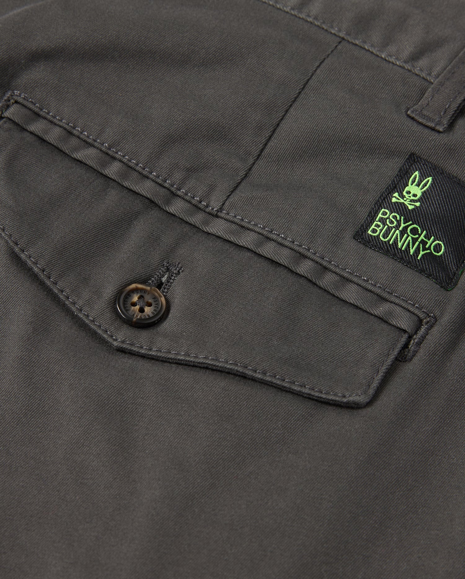 Detailed view of the gray fabric featuring a buttoned pocket, designed for stretch and comfort. Positioned above the pocket is a green and black label showcasing a bunny logo along with "Psycho Bunny," evoking the classic style of the MENS GILMAN CHINO PANT - B6P393Z1WB.