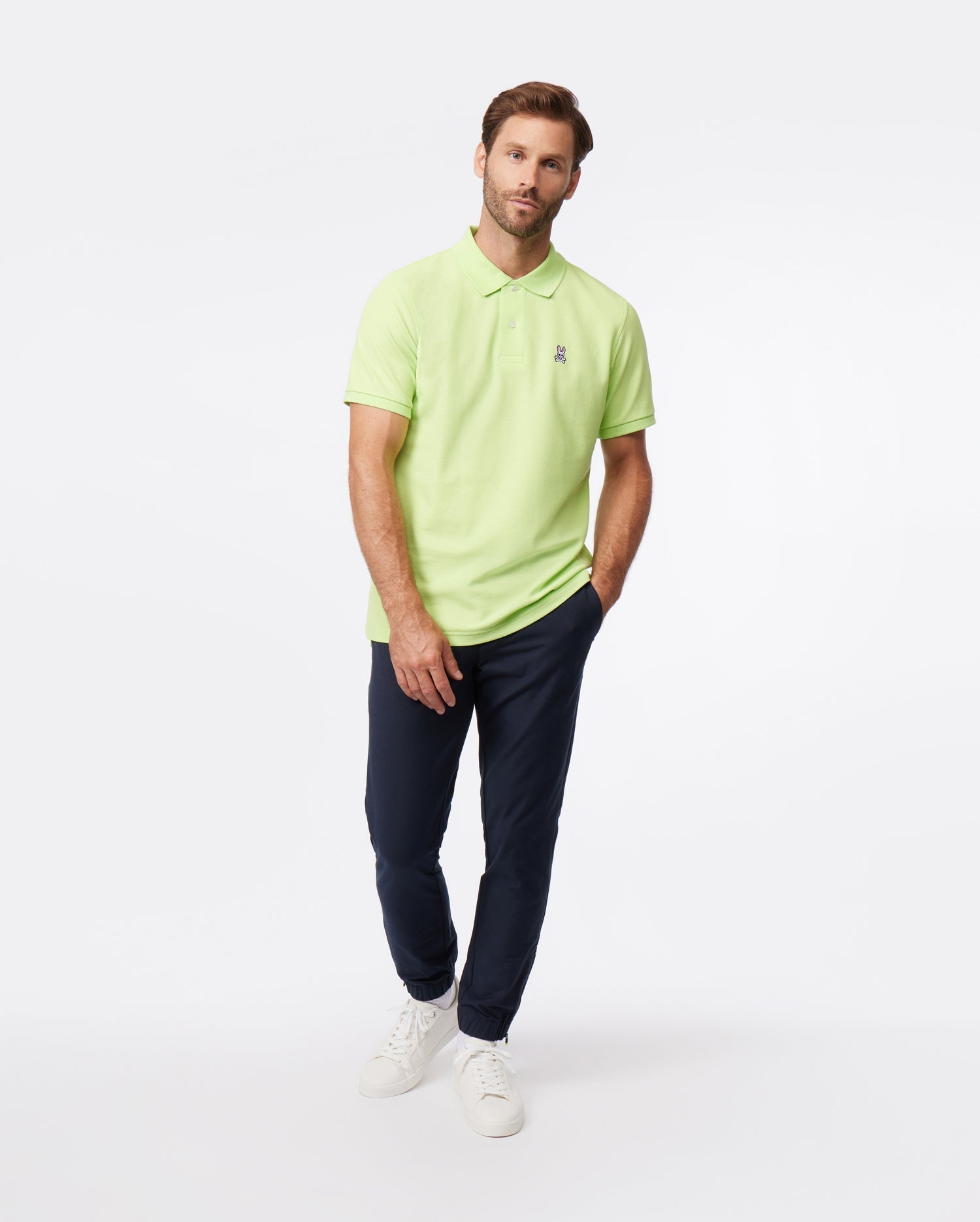 A man stands against a white background, wearing a lime green polo shirt, Psycho Bunny's MENS MADISON COMMUTER PANT - B6P395Z1WB with an elasticized waistband, and white sneakers, looking at the camera with a neutral expression.