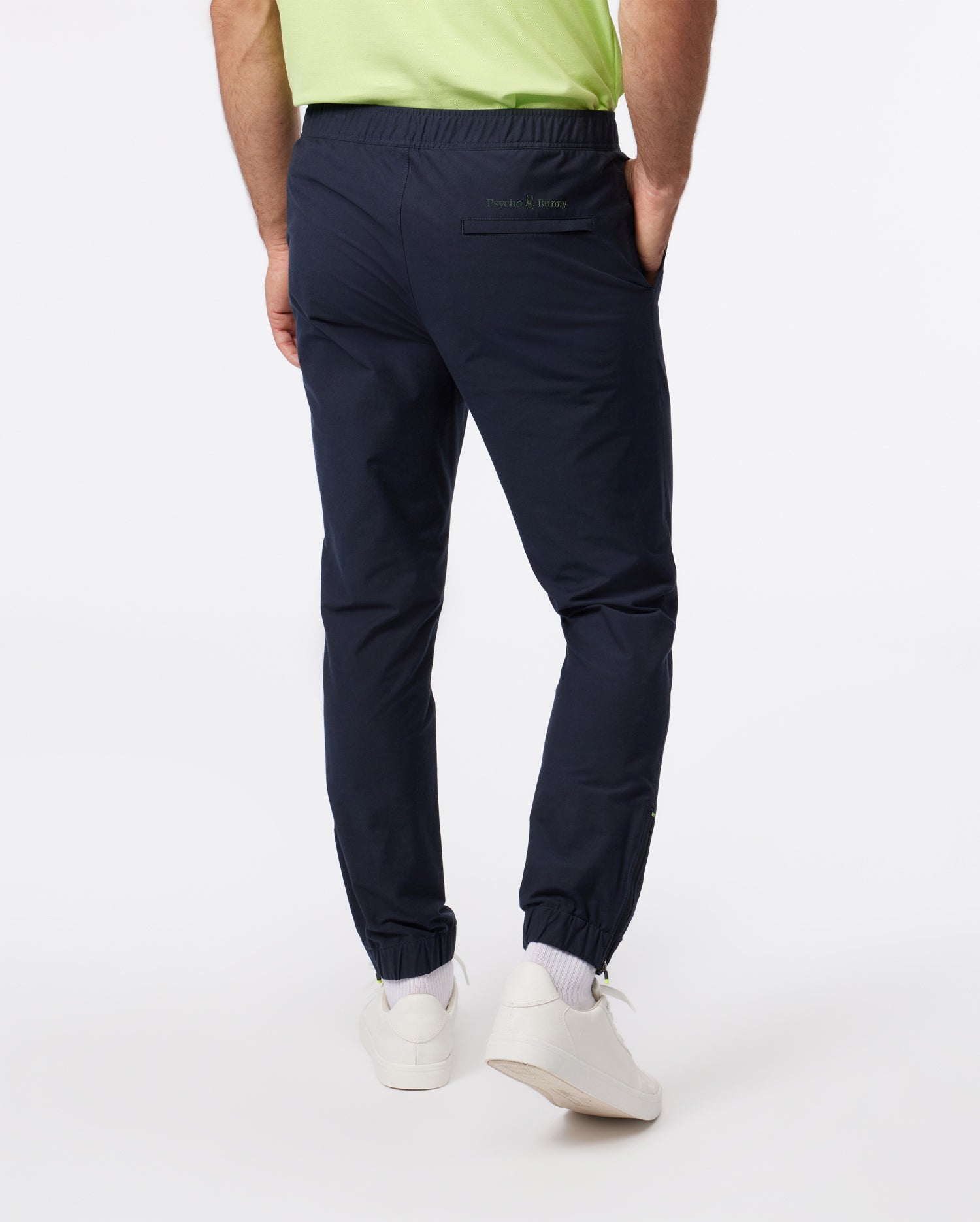 Rear view of a model wearing Psycho Bunny's MEN'S MADISON COMMUTER PANT - B6P395Z1WB joggers with an elasticized waistband, paired with a green t-shirt, standing against a white background. The model's footwear includes casual white sneakers.