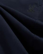 Close-up image of dark blue stretch woven material with subtle folds, featuring a small, embroidered green Psycho Bunny character in the lower left corner.