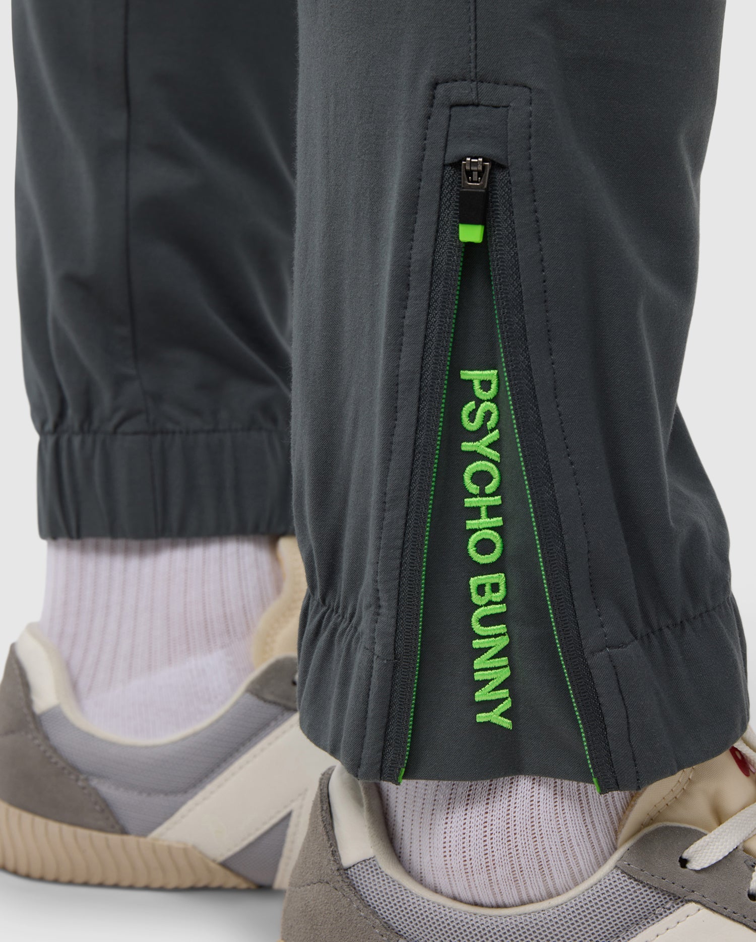 Close-up of a person's lower leg showing the embroidered Psycho Bunny logos on black MENS MADISON COMMUTER PANT - B6P395Z1WB with the text "psycho bunny" in green and a pair of grey sneakers.