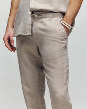 A person wears Psycho Bunny’s MENS FATE LINEN DRAWSTRING PANT (B6P471C200) in light beige linen blend, paired with a matching button-up shirt and a beige beaded bracelet. They stand with one hand in their pocket against a neutral background.