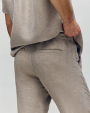 A person models the Psycho Bunny MENS FATE LINEN DRAWSTRING PANT (B6P471C200) in beige, highlighting the back pocket and textured linen blend for cooling comfort. Their left hand rests on their hip against a plain, neutral background.