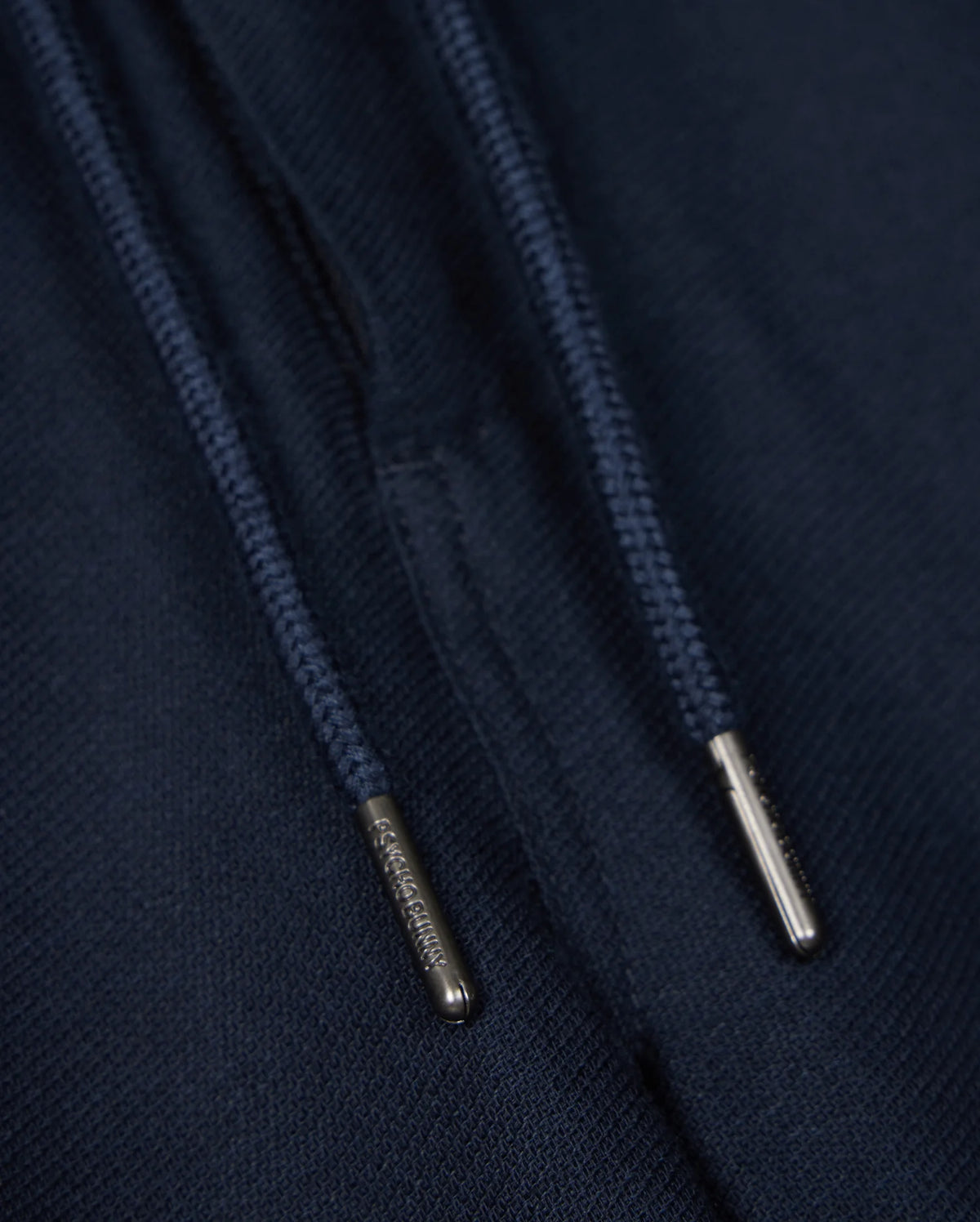 Close-up of navy blue drawstrings with silver metal tips on a dark fabric background, perfect for the Psycho Bunny MENS FATE LINEN DRAWSTRING PANT - B6P471C200. The metal tips have small engraved text, but the content is not fully readable in the image. Ideal for those summer trousers that need both style and durability.