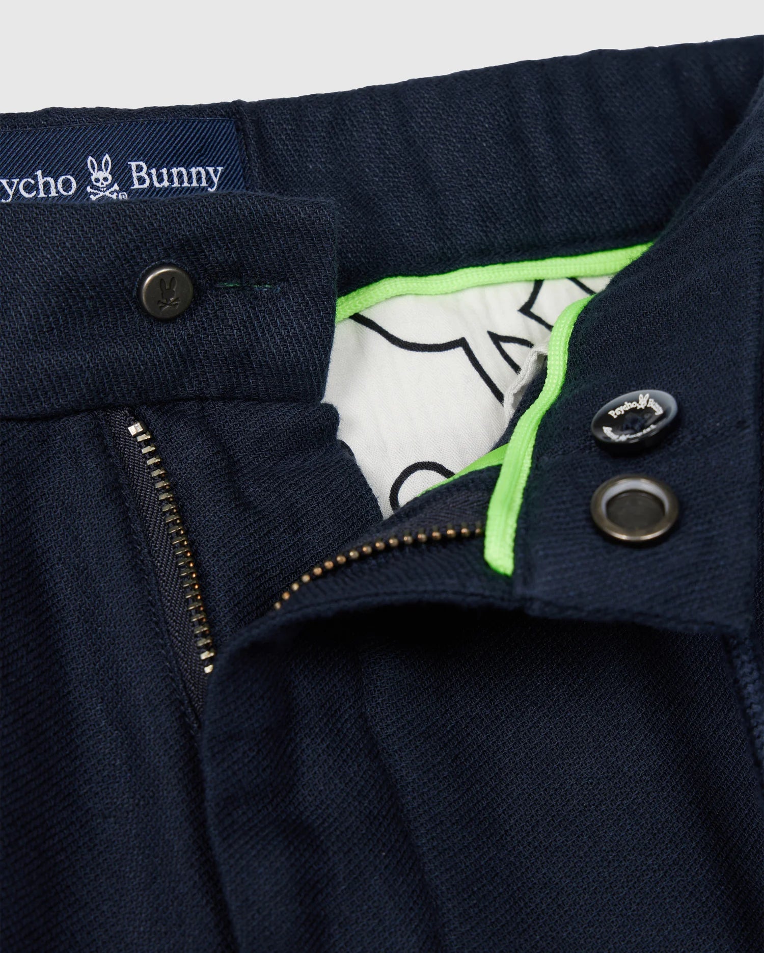 Close-up of the waistband and front fly of a pair of Men's Fate Linen Drawstring Pants in dark blue, featuring a partial zip and two dark buttons. The inner lining is white with a black line illustration and neon green trim. A label inside reads "Psycho Bunny" alongside a small bunny logo.