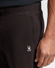 A person in dark-colored Psycho Bunny MENS LEVY ESSENTIAL FLEECE SWEATPANT, featuring a small white rabbit skull and crossbones logo on the side. Their right hand rests partially visible near the pocket.