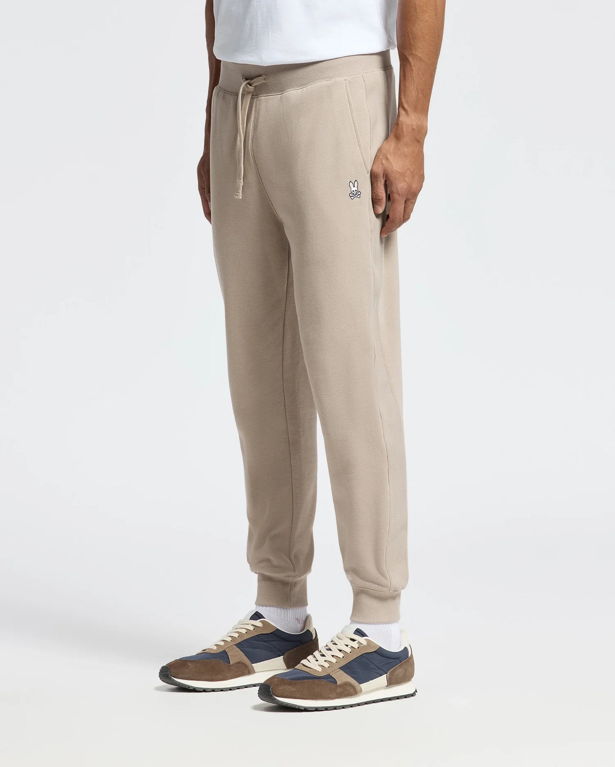 A person is dressed in Psycho Bunny's MENS LEVY ESSENTIAL FLEECE SWEATPANT, featuring a small embroidered logo on the upper thigh, paired with a white top and brown and navy sneakers. The spotlight is on these cozy cotton sweatpants against a plain white background.