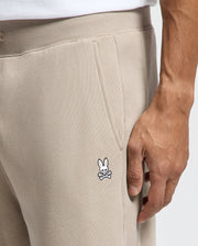 A person is wearing the Psycho Bunny MENS LEVY ESSENTIAL FLEECE SWEATPANT, featuring a discreet embroidered bunny and crossbones logo on the pocket. They are standing with their hand resting near the logo, while a white shirt peeks out from above.