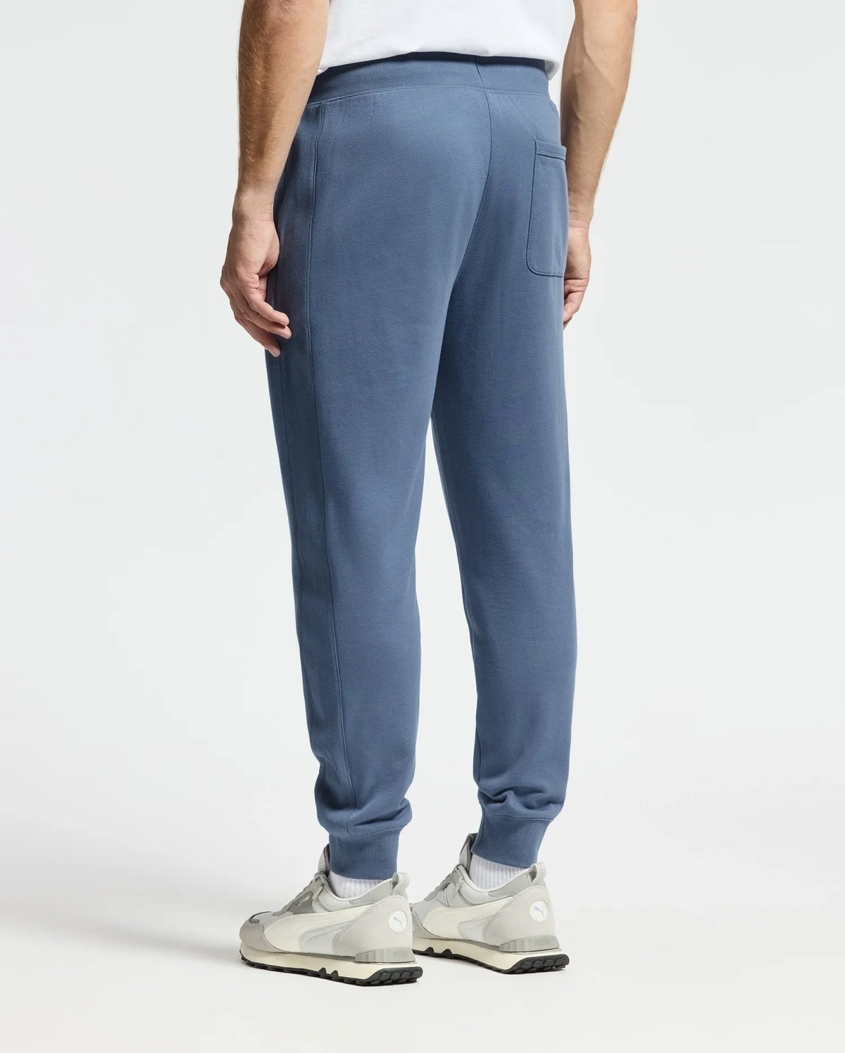 A person stands wearing light blue, Psycho Bunny's MENS LEVY ESSENTIAL FLEECE SWEATPANT and white sneakers. The image captures their back from the waist down, set against a plain white background.