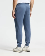 A person stands wearing light blue, Psycho Bunny's MENS LEVY ESSENTIAL FLEECE SWEATPANT and white sneakers. The image captures their back from the waist down, set against a plain white background.