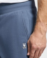 A close-up of someone wearing the Psycho Bunny MENS LEVY ESSENTIAL FLEECE SWEATPANT - B6P721D200, featuring a small embroidered rabbit skull and crossbones logo near the pocket. The person's hand rests on the comfortable blue cotton fabric of the pants.