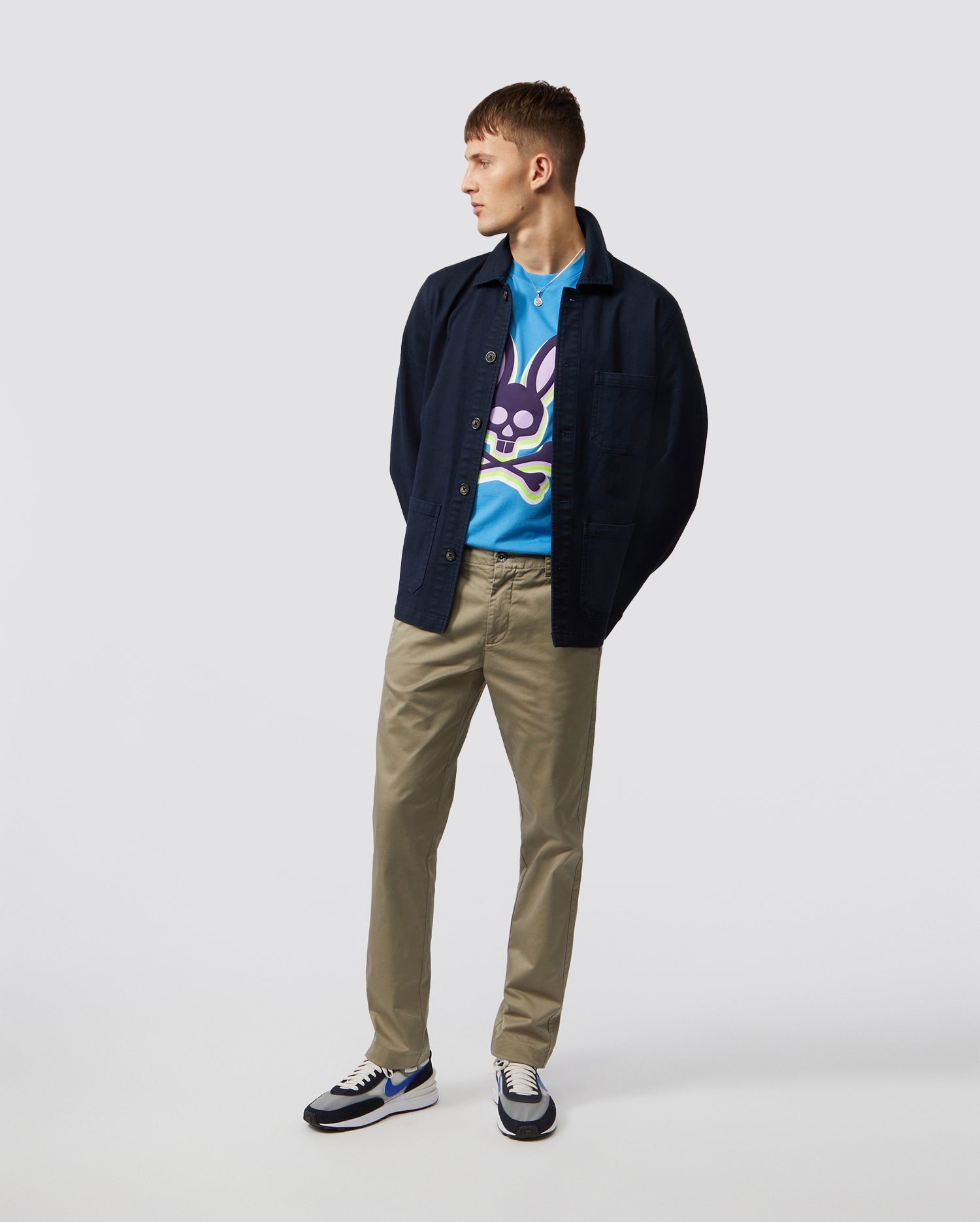 A person stands against a plain background wearing a blue graphic t-shirt with a purple character, Psycho Bunny's MENS DELANCY CHINO PANTS in beige stretch cotton, and a dark jacket. White sneakers with blue detailing complete the look as they gaze to the side.