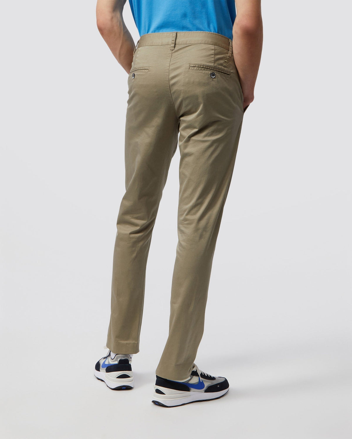 Rear view of a person wearing Psycho Bunny's MEN'S DELANCY CHINO PANTS - B6P784U1CO, a blue shirt, and white sneakers with black and blue accents against a plain light-colored background.