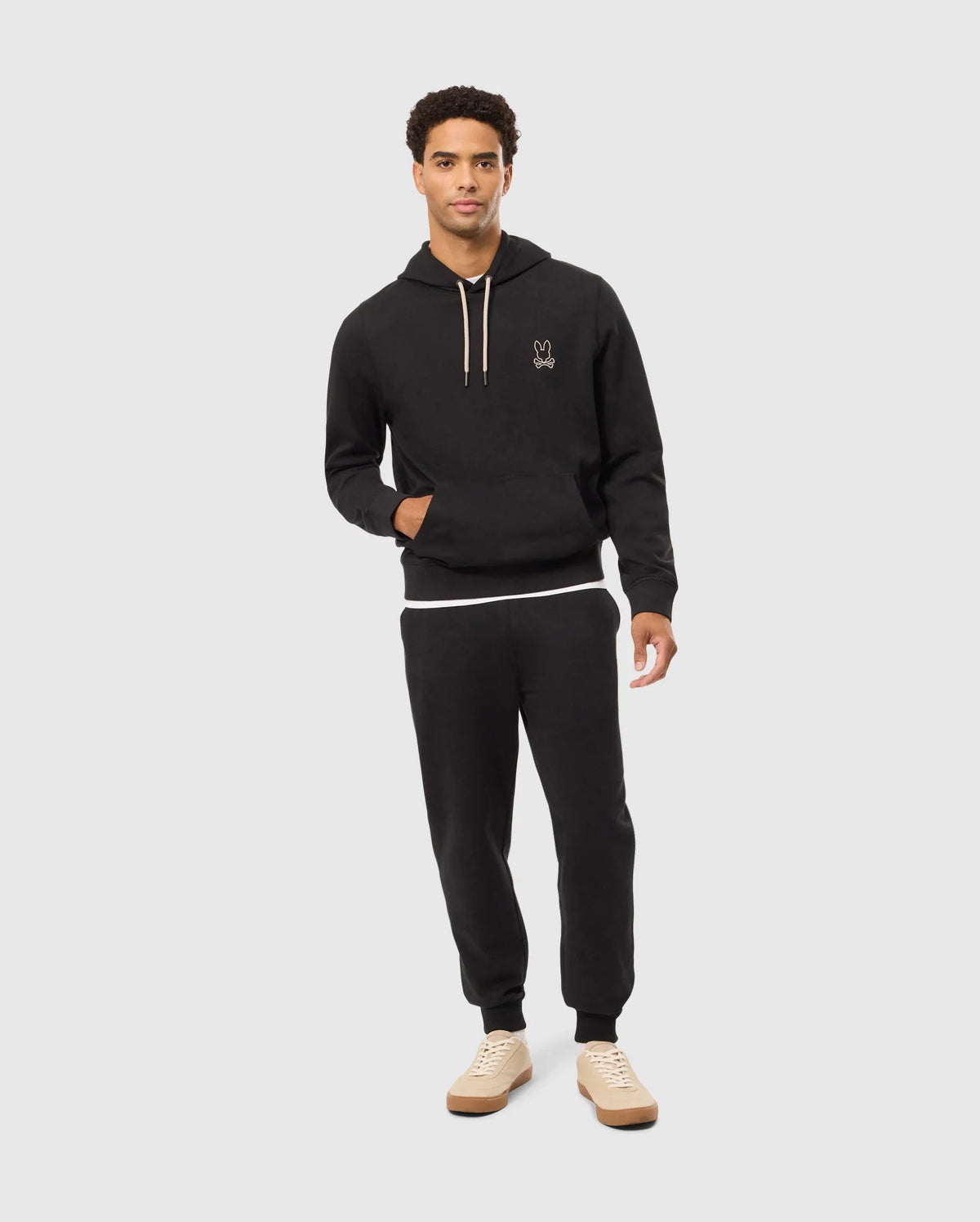 A person is wearing a Psycho Bunny black cotton-blend hoodie paired with the MENS JAMES PREMIUM SWEATPANT - B6P791D200. They have on beige sneakers, and the hoodie showcases a minimalistic design with a small logo on the left chest. The individual is standing against a plain light gray background.