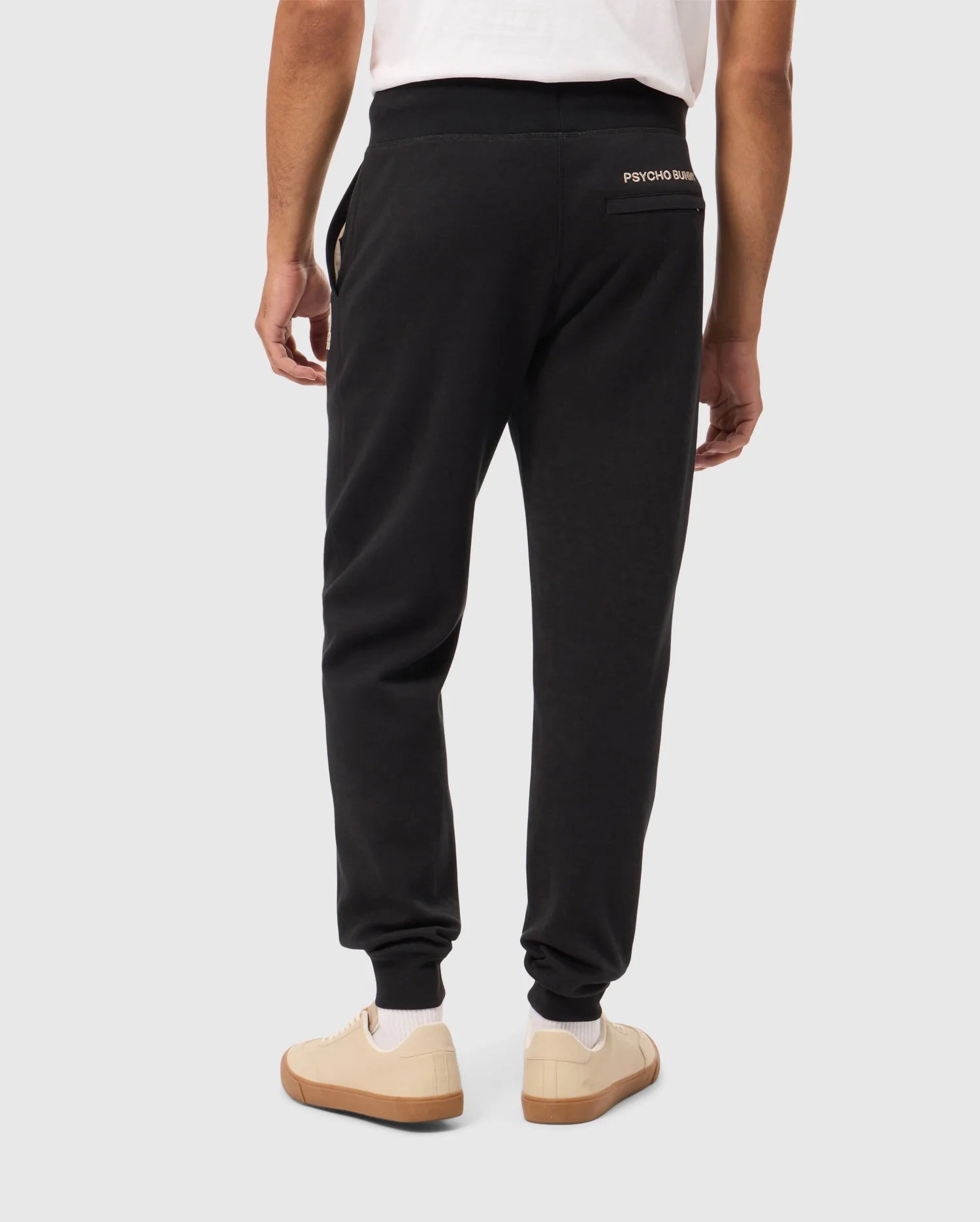 Rear view of a person wearing Psycho Bunny's James Premium Sweatpant in black cotton-blend and a white t-shirt. They stand against a plain background, showcasing white sneakers with tan soles. These premium sweatpants feature the distinctive small logo on the back, near the waistband.