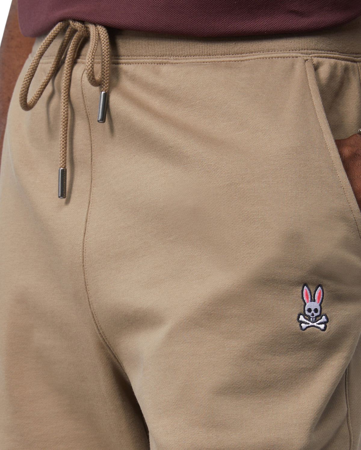 Close-up of a person wearing Psycho Bunny's Men's French Terry Knit Sweatpants in classic fit khaki with a drawstring waist, featuring an embroidered pink-eared bunny and crossbones logo near the pocket. The person has tucked a dark shirt into the pants.