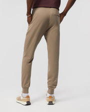 A person wearing Psycho Bunny's men's French terry knit sweatpants (B6P828ARFT) and white sneakers with beige accents stands with their hands in their pockets, showcasing the relaxed fit and back pocket of the joggers, set against a plain white background.