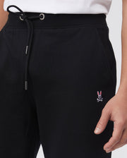A person is seen wearing Psycho Bunny's black MENS FRENCH TERRY KNIT SWEATPANTS - B6P828ARFT, featuring a classic fit with a small pink bunny and crossbones embroidery on the upper leg. The drawstring waist is paired with a white top, revealing only the lower torso and part of an arm.