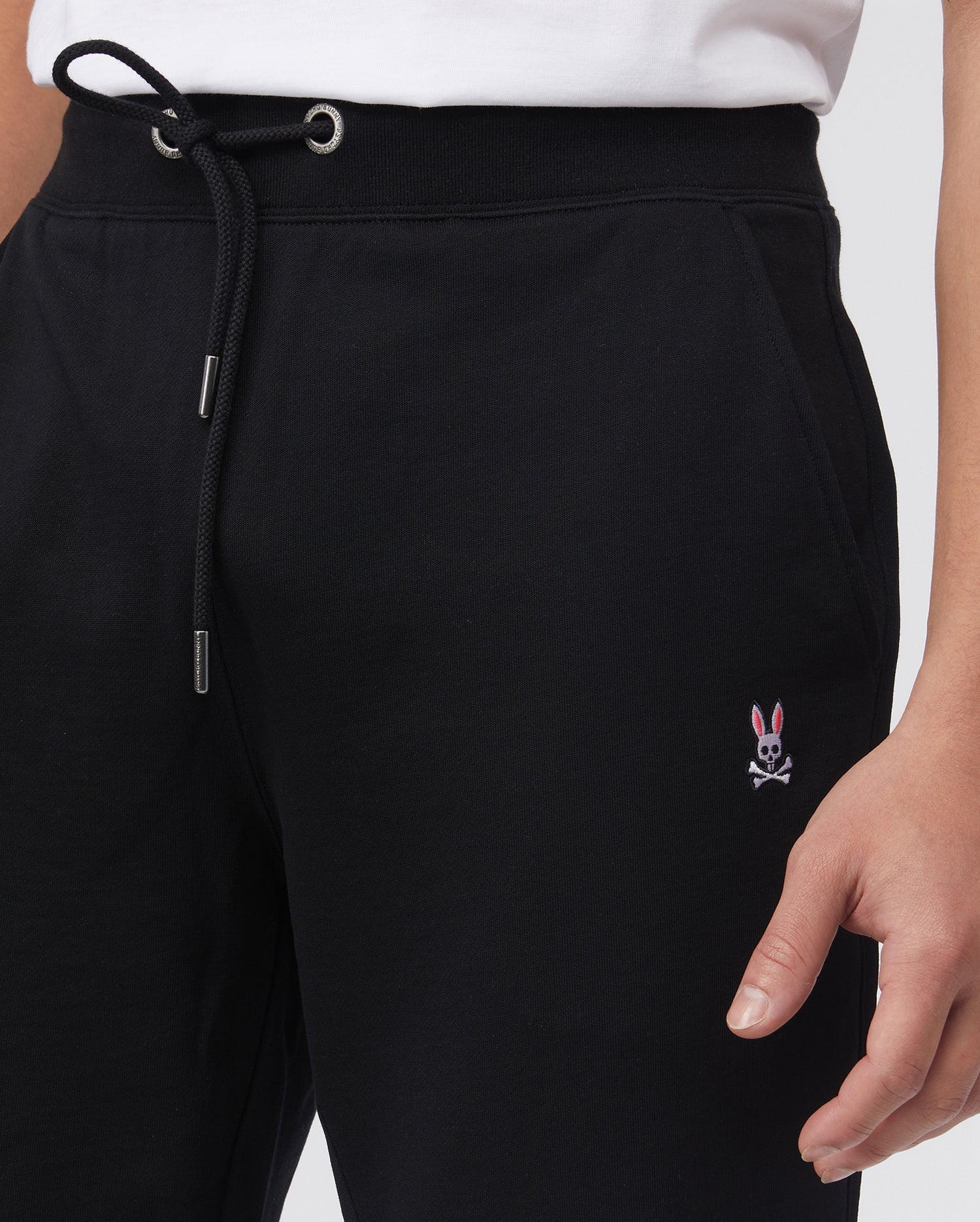 A person is seen wearing Psycho Bunny's black MENS FRENCH TERRY KNIT SWEATPANTS - B6P828ARFT, featuring a classic fit with a small pink bunny and crossbones embroidery on the upper leg. The drawstring waist is paired with a white top, revealing only the lower torso and part of an arm.