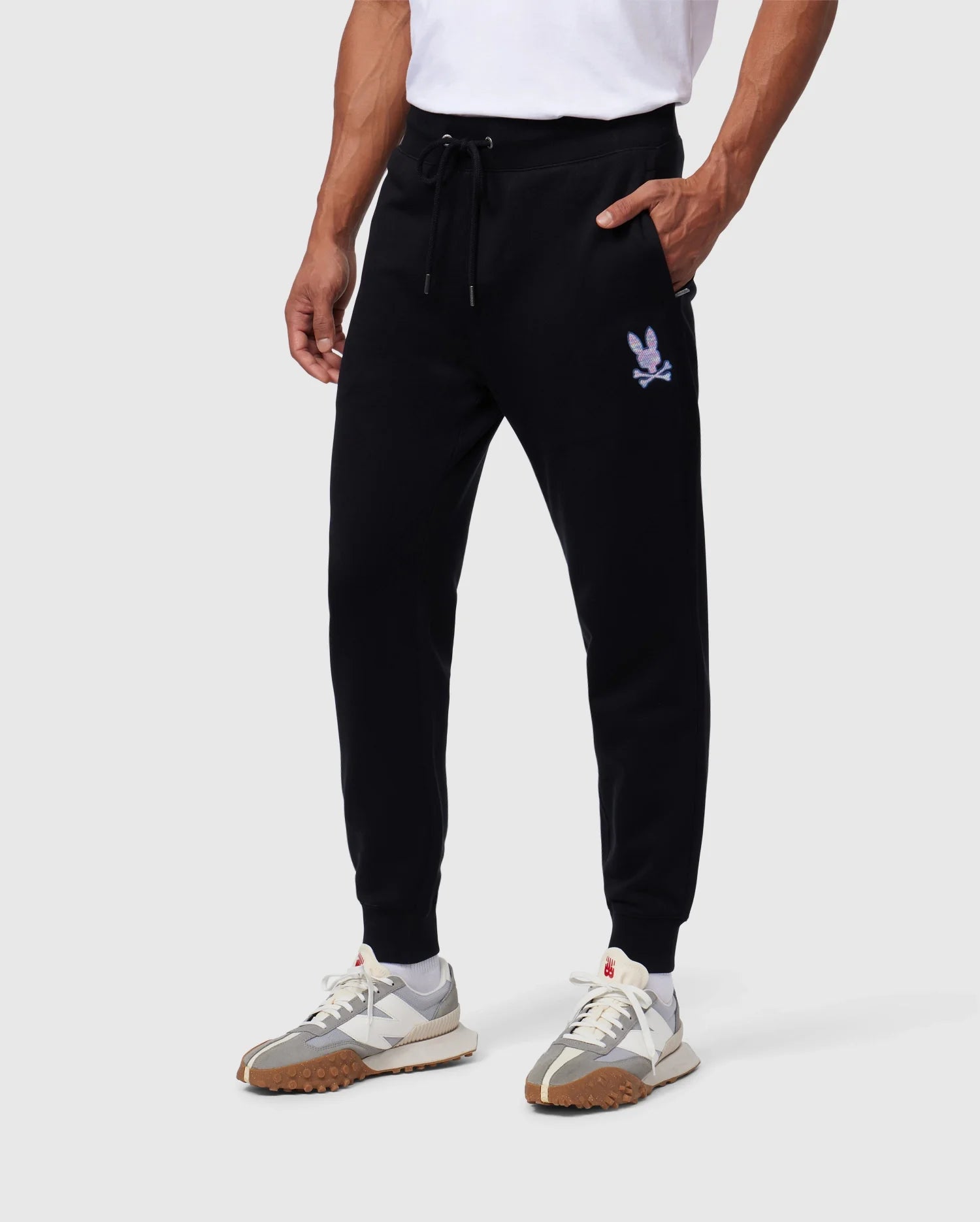 A person wearing black Psycho Bunny MENS SANTA CRUZ SWEATPANT - B6P843A2FT with a small, white embroidered logo on the left thigh. The joggers, made from 100% cotton, have an elastic waistband with a drawstring for both comfort and style. With their hands in the pockets, they also sport white sneakers with grey and brown accents.