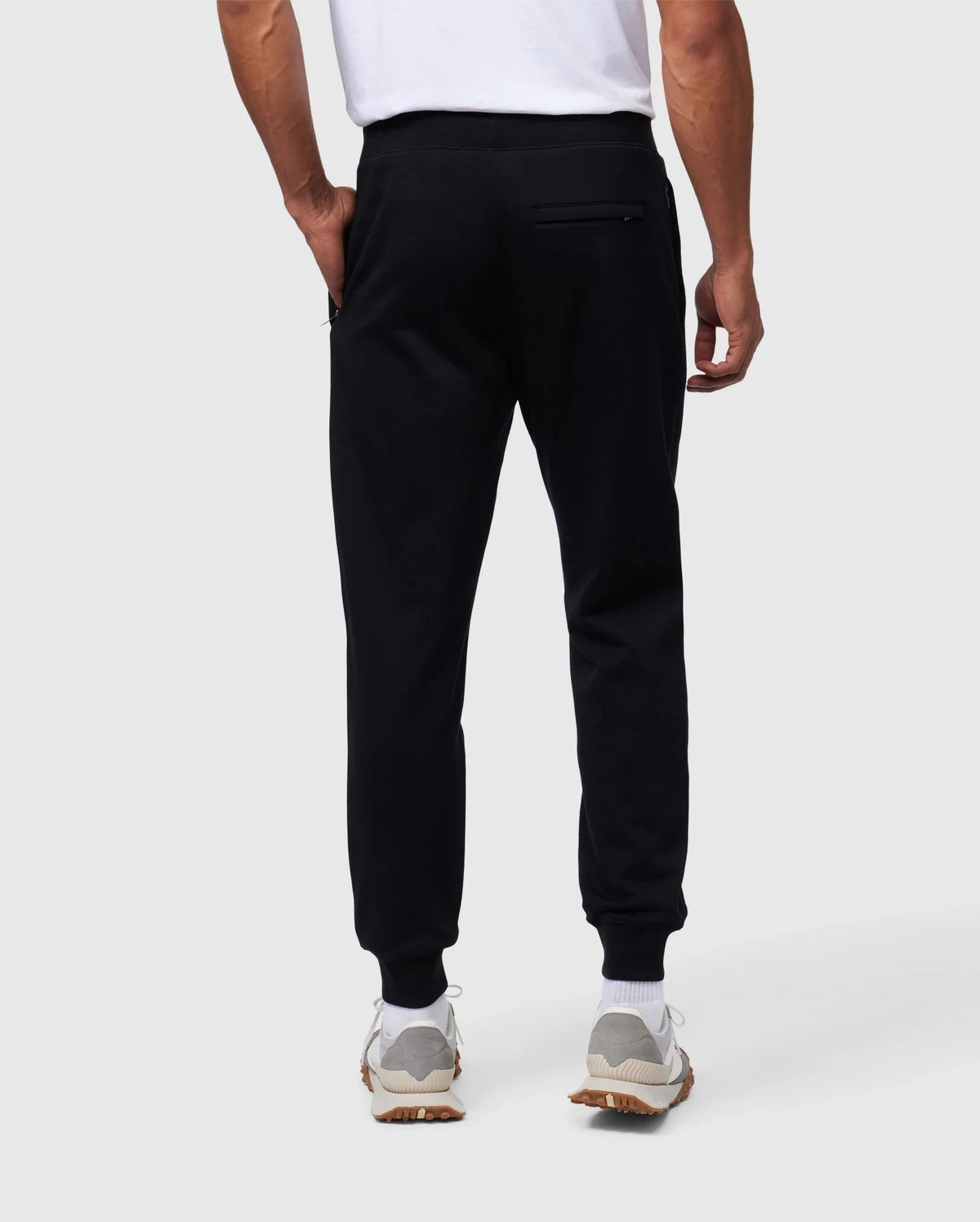 Back view of a person wearing Psycho Bunny MENS SANTA CRUZ SWEATPANT - B6P843A2FT, a white t-shirt, and white sneakers. The 100% cotton sweatpants feature a zippered pocket on the right side for added comfort and style. The person stands with their left hand in a pocket against a plain, light-colored background.