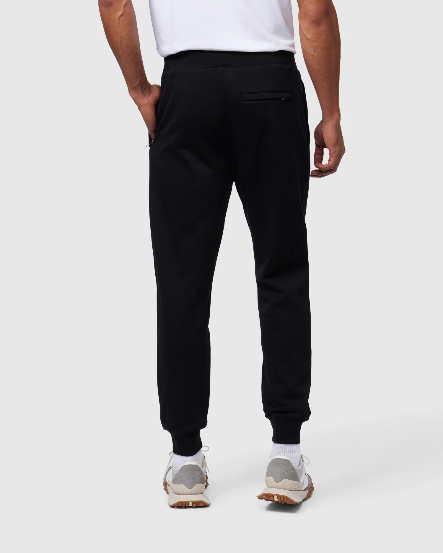 Back view of a person wearing Psycho Bunny MENS SANTA CRUZ SWEATPANT - B6P843A2FT, a white t-shirt, and white sneakers. The 100% cotton sweatpants feature a zippered pocket on the right side for added comfort and style. The person stands with their left hand in a pocket against a plain, light-colored background.
