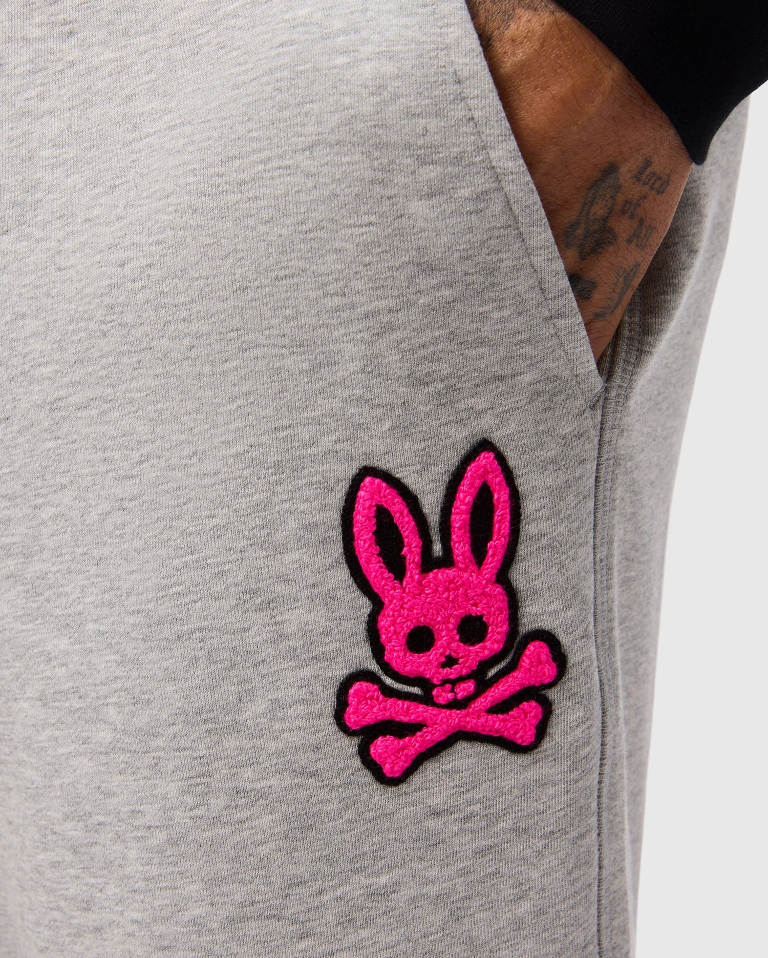 Close-up of someone wearing Psycho Bunny's SCOTT CHENILLE PATCH SWEATPANT - B6P856D200, featuring a bright pink bunny skull and crossbones patch, with a tattooed hand partially visible.