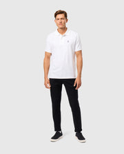 A man wearing a regular fit white polo shirt and the MENS HAYS TWILL PANT - B6P894D200 from Psycho Bunny stands against a plain gray background. His cotton-blend twill outfit is paired with black sneakers, offering both style and comfort.