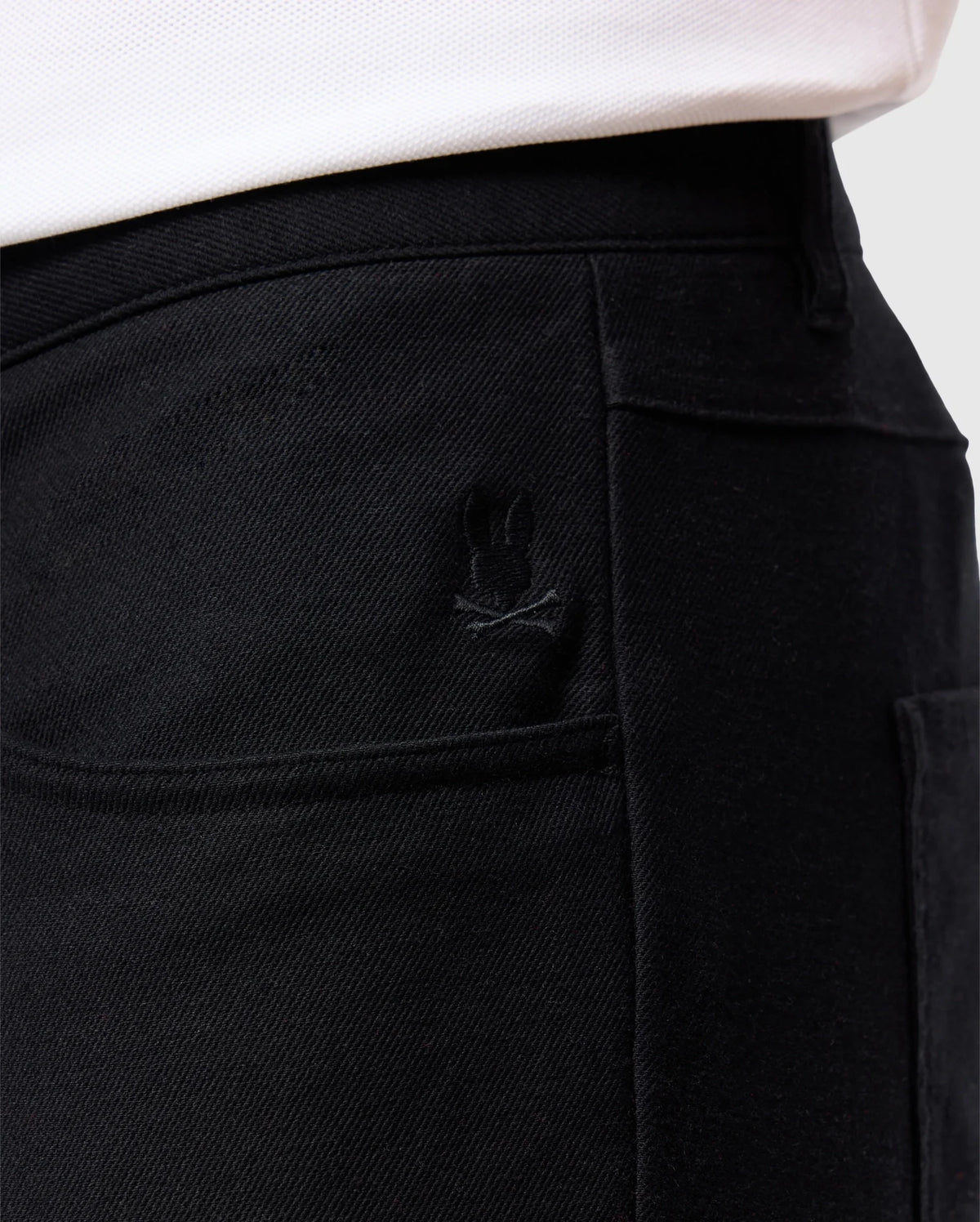 Close-up of a person wearing the Psycho Bunny MENS HAYS TWILL PANT - B6P894D200 in a regular fit and black color, made from cotton-blend twill, featuring a stitched design above one of the back pockets. The edge of a white shirt is visible at the top.