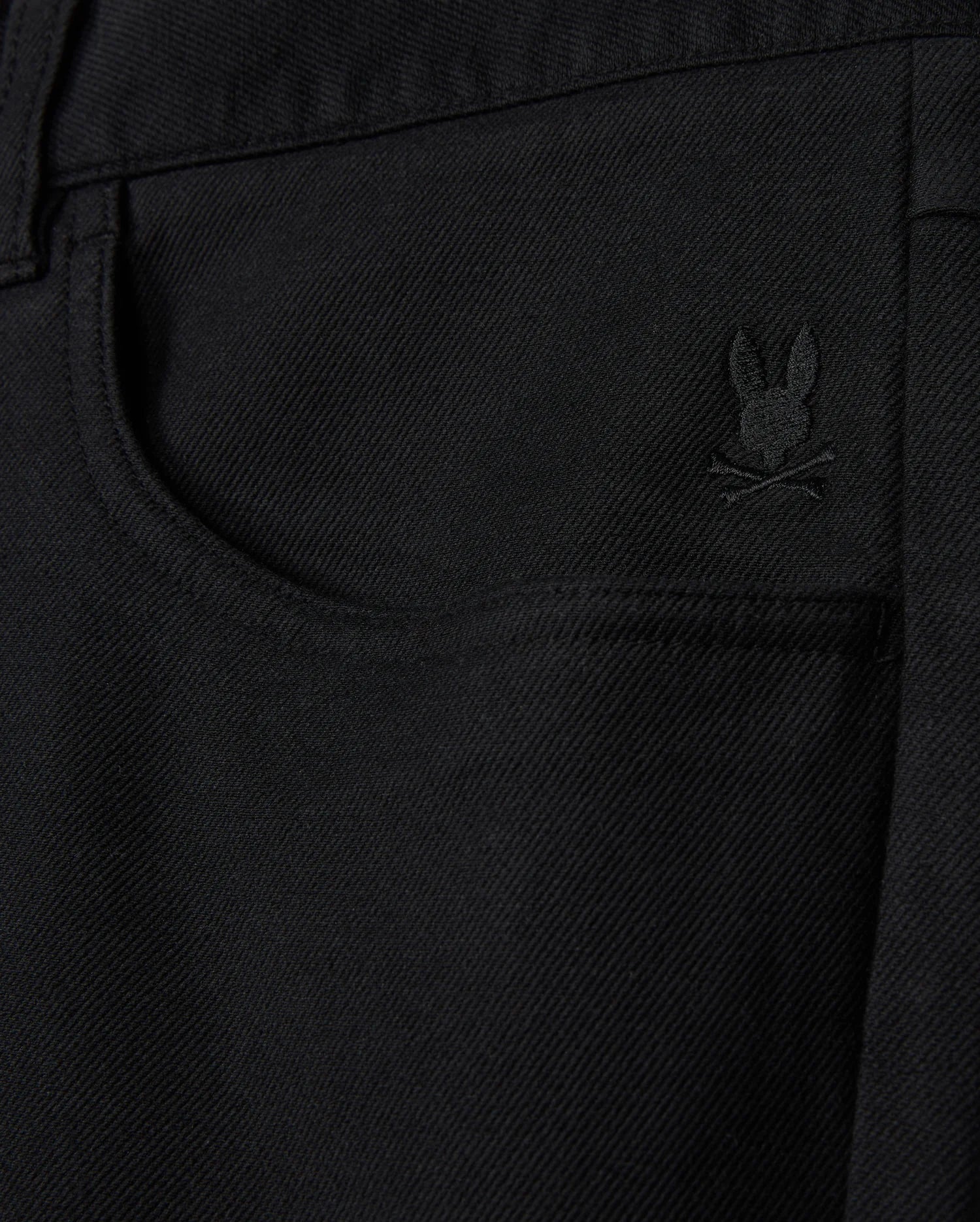 Close-up of a black denim pocket on the Psycho Bunny MENS HAYS TWILL PANT - B6P894D200, featuring an embroidered rabbit's head with crossed bones beneath it. This subtle design detail enhances this regular fit piece, crafted from a cotton-blend twill.