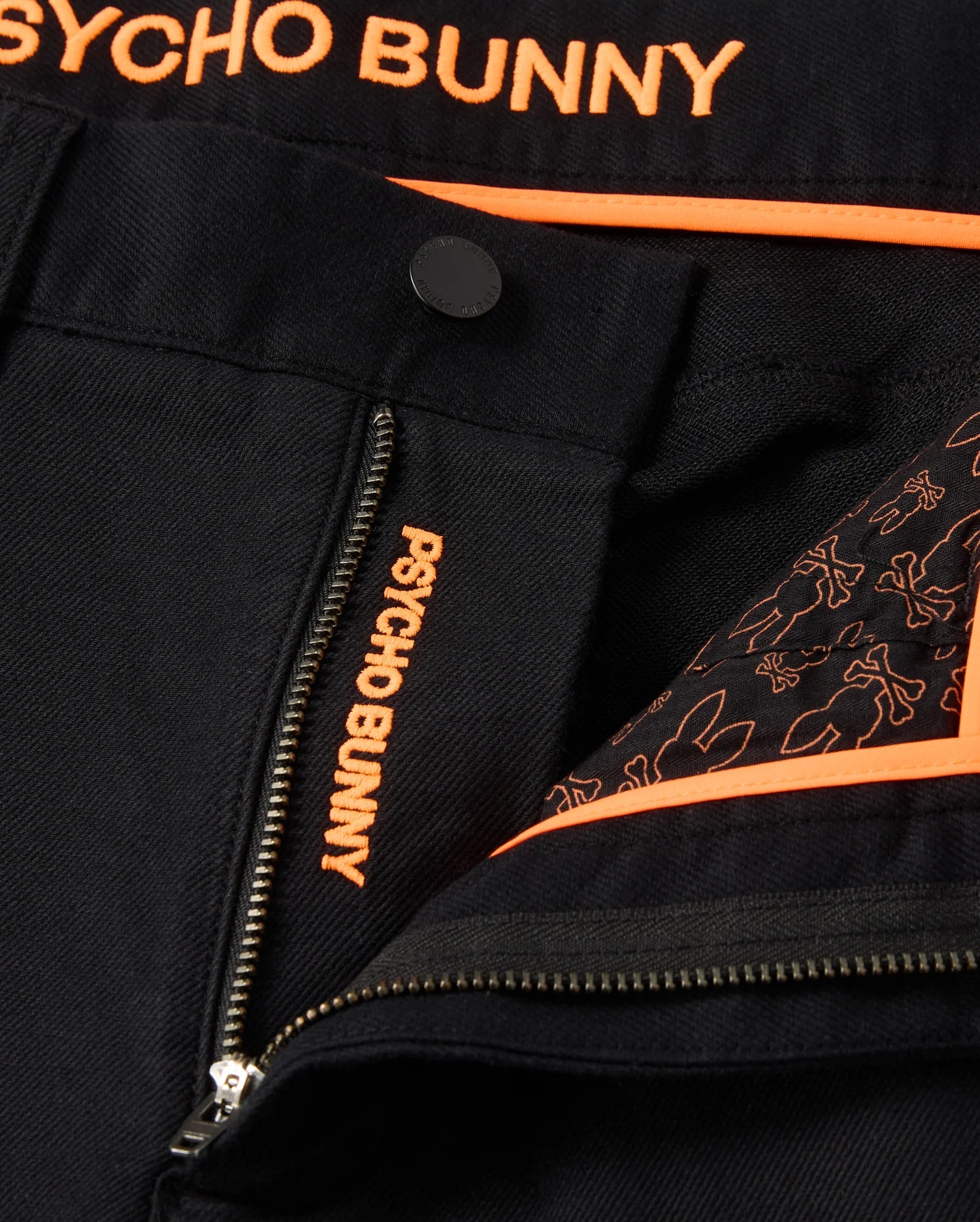 A close-up shot showcases the MENS HAYS TWILL PANT by Psycho Bunny in black, adorned with orange embroidery spelling "Psycho Bunny." Made from a cotton-blend twill with four-way stretch for enhanced comfort, these pants feature a regular fit. A partially open zipper reveals a lining decorated with an orange bunny pattern, complemented by orange trim and detailing.