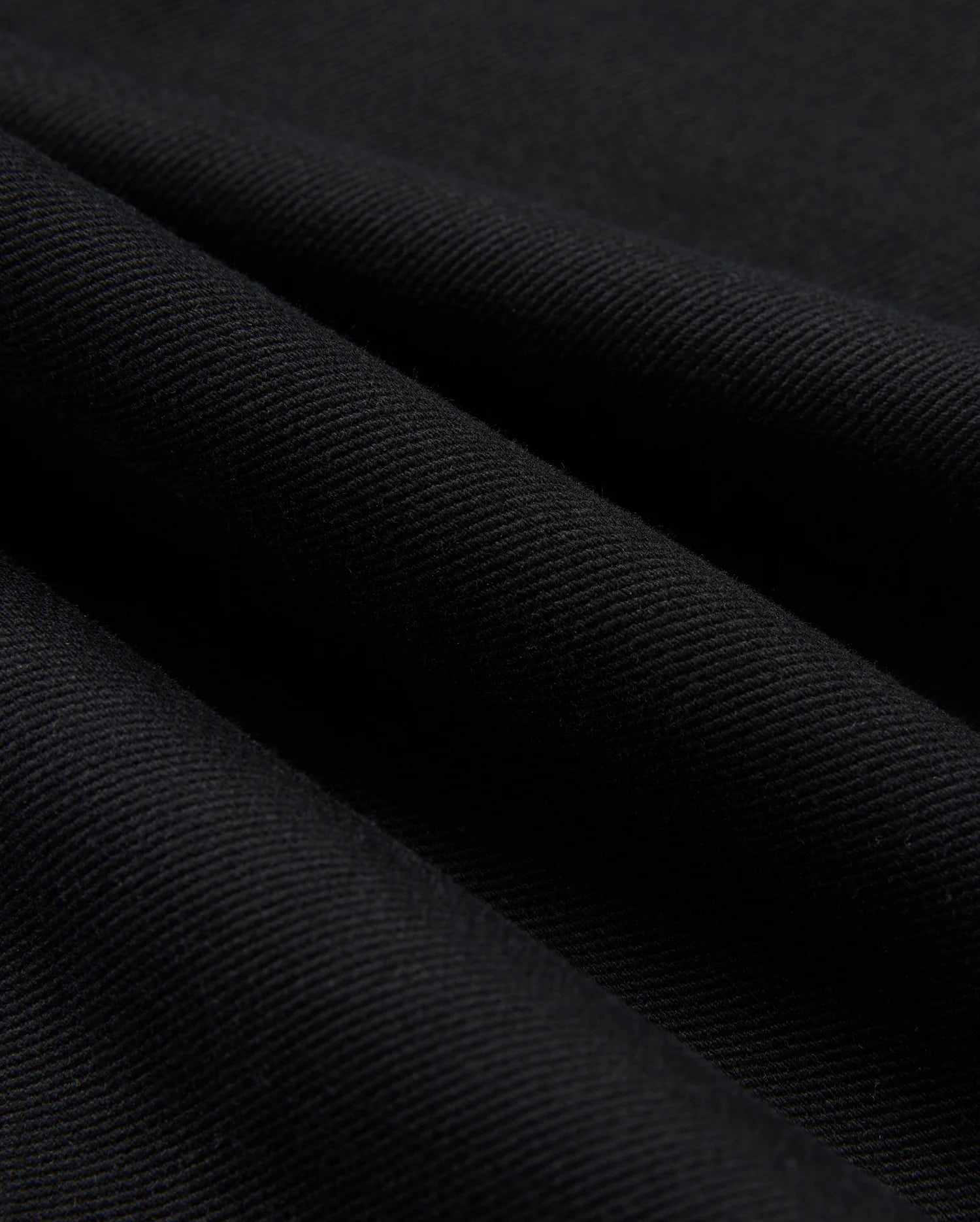 Close-up of Psycho Bunny's MENS HAYS TWILL PANT - B6P894D200, crafted from a black cotton-blend twill fabric with a diagonal ribbed texture. The material boasts four-way stretch, giving it a soft feel and a subtle sheen, with folds producing shadowy areas across the surface.