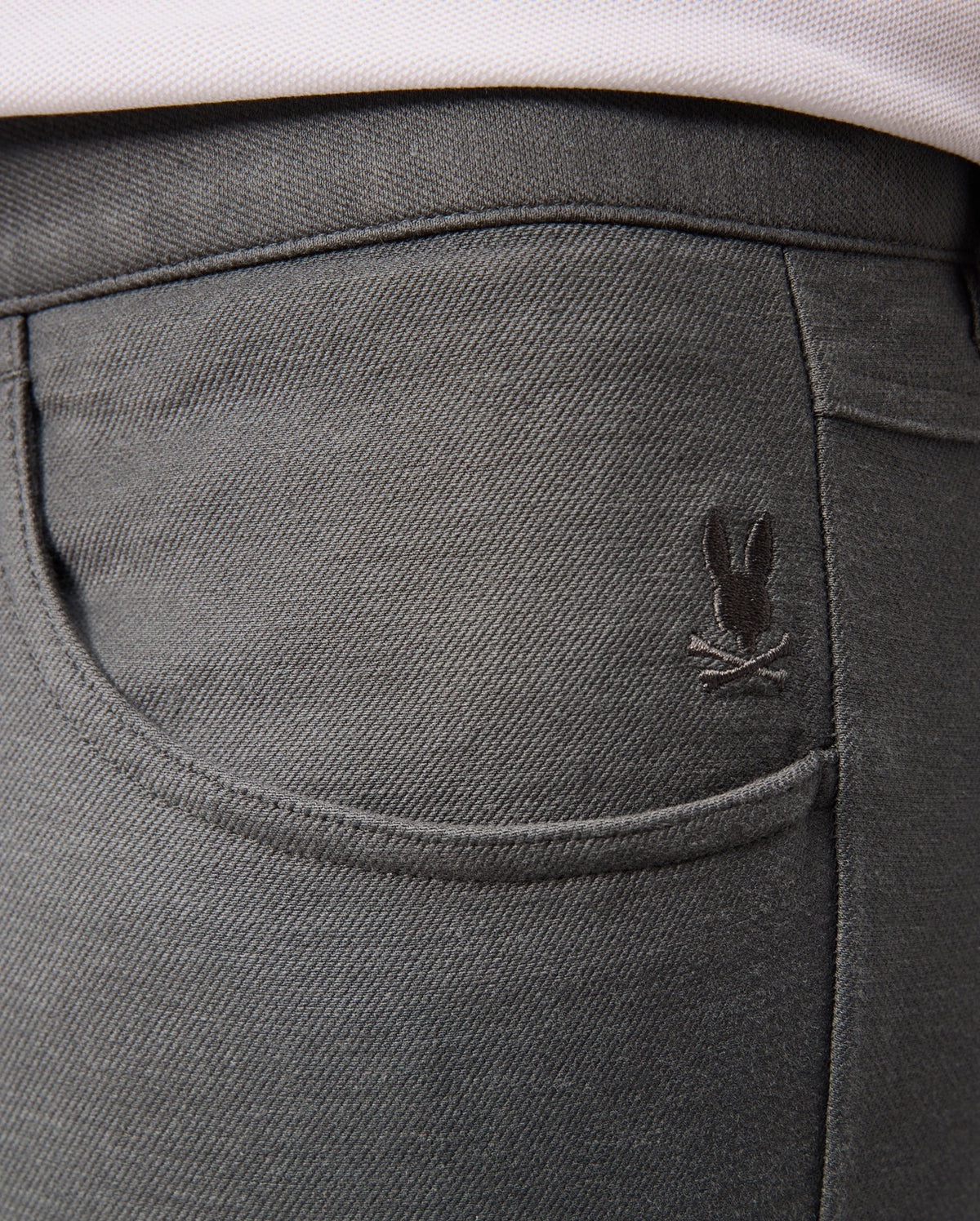 A close-up of the back pocket of the Psycho Bunny MENS HAYS TWILL PANT - B6P894D200 in dark gray cotton-blend twill showcases a small embroidered design featuring a bunny with crossed bones above the pocket. The fabric provides 4-way stretch and has a slightly textured appearance.