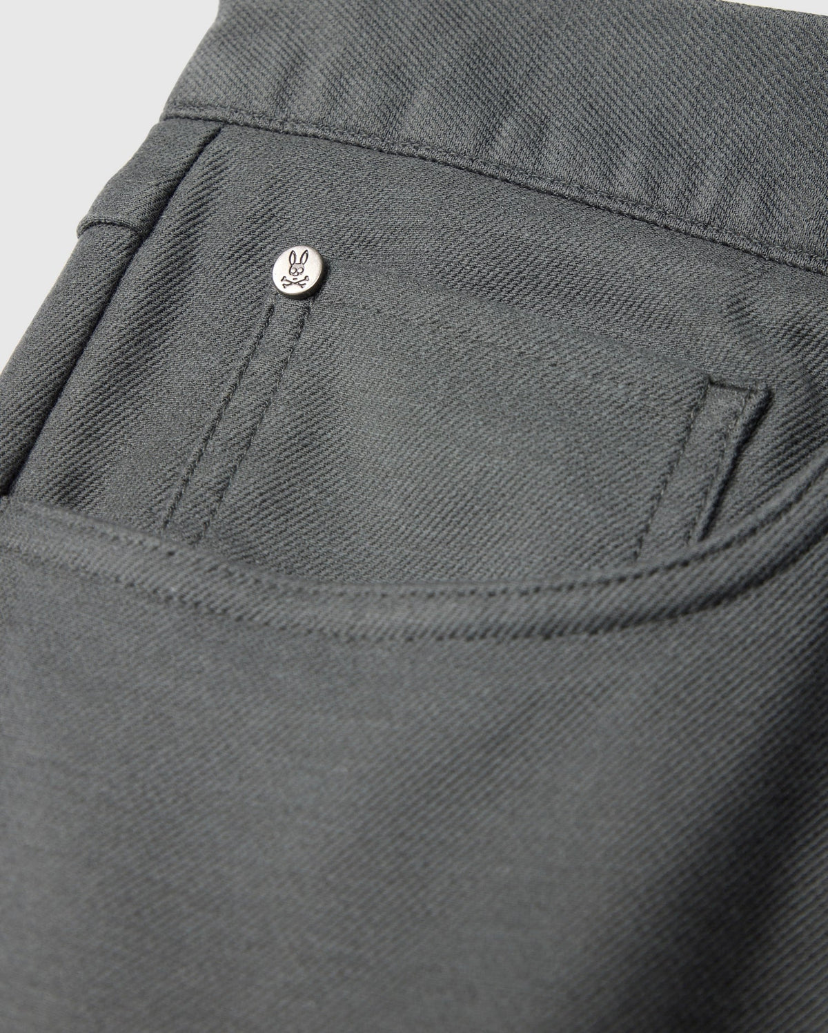 Close-up of the MENS HAYS TWILL PANT - B6P894D200 by Psycho Bunny, showcasing a pocket adorned with a small metallic button engraved with a minimalist face design. Situated on the pocket's upper corner, this dark gray cotton-blend twill garment exhibits 4-way stretch and features a textured, sturdy fabric.