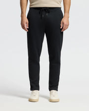 A person wearing black Men's Monroe Twill Patch Fleece Sweatpants by Psycho Bunny and white sneakers stands against a gray background. Their hands rest naturally by their sides, and the drawstring of the 100% cotton pants is tied in a bow at the waist.