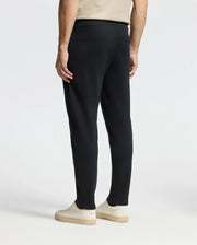 A person in a beige t-shirt and the Psycho Bunny MENS MONROE TWILL PATCH FLEECE SWEATPANT is seen from behind, showcasing black sweatpants crafted from 100% cotton. They are wearing white sneakers against a plain, minimalistic background.