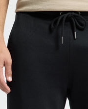 A close-up of a person wearing Psycho Bunny's MENS MONROE TWILL PATCH FLEECE SWEATPANT (B6P929D200) paired with a beige shirt. The drawstrings are tied in a bow at the waistband, while the left hand rests relaxed at their side against a neutral background.
