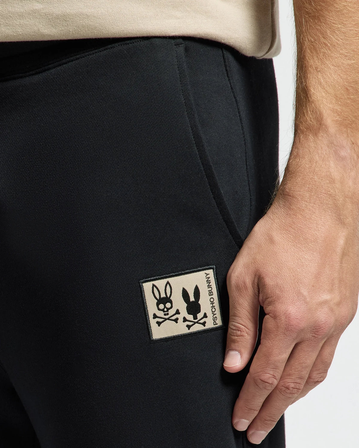 A close-up shot shows someone wearing the MENS MONROE TWILL PATCH FLEECE SWEATPANT by Psycho Bunny in black. One hand subtly rests near the pocket, where the iconic logo—a stylized bunny with crossbones underneath and "Psycho Bunny" branding—is prominently displayed.