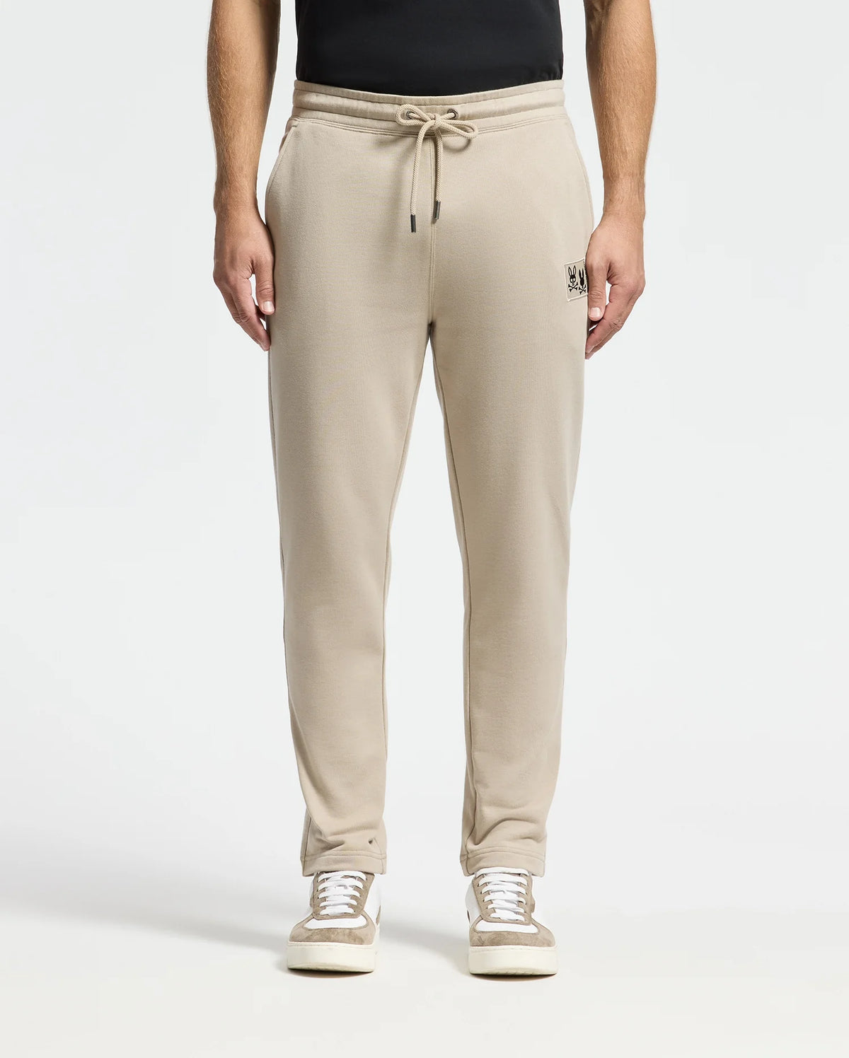 A person wearing the Psycho Bunny MENS MONROE TWILL PATCH FLEECE SWEATPANT - B6P929D200, designed in beige with a soft French terry texture and a subtle embroidered logo on the left pocket, stands against a plain background. The pants are made from 100% cotton, featuring a drawstring waistband for a relaxed fit. They are paired with casual sneakers and a black top.
