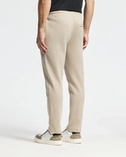 A person dressed in beige Monroe Twill Patch Fleece Sweatpants by Psycho Bunny and white sneakers with brown accents is standing against a plain white background. The image focuses on the lower half of the body from behind, showcasing the casual elegance of 100% cotton comfort.
