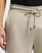 A person wearing the Psycho Bunny beige MENS MONROE TWILL PATCH FLEECE SWEATPANT stands against a plain background. With their left hand hanging by their side, the sweatpants' waistband displays a visible seam. These sweatpants, made of 100% cotton, are paired with a black top for a classic look.