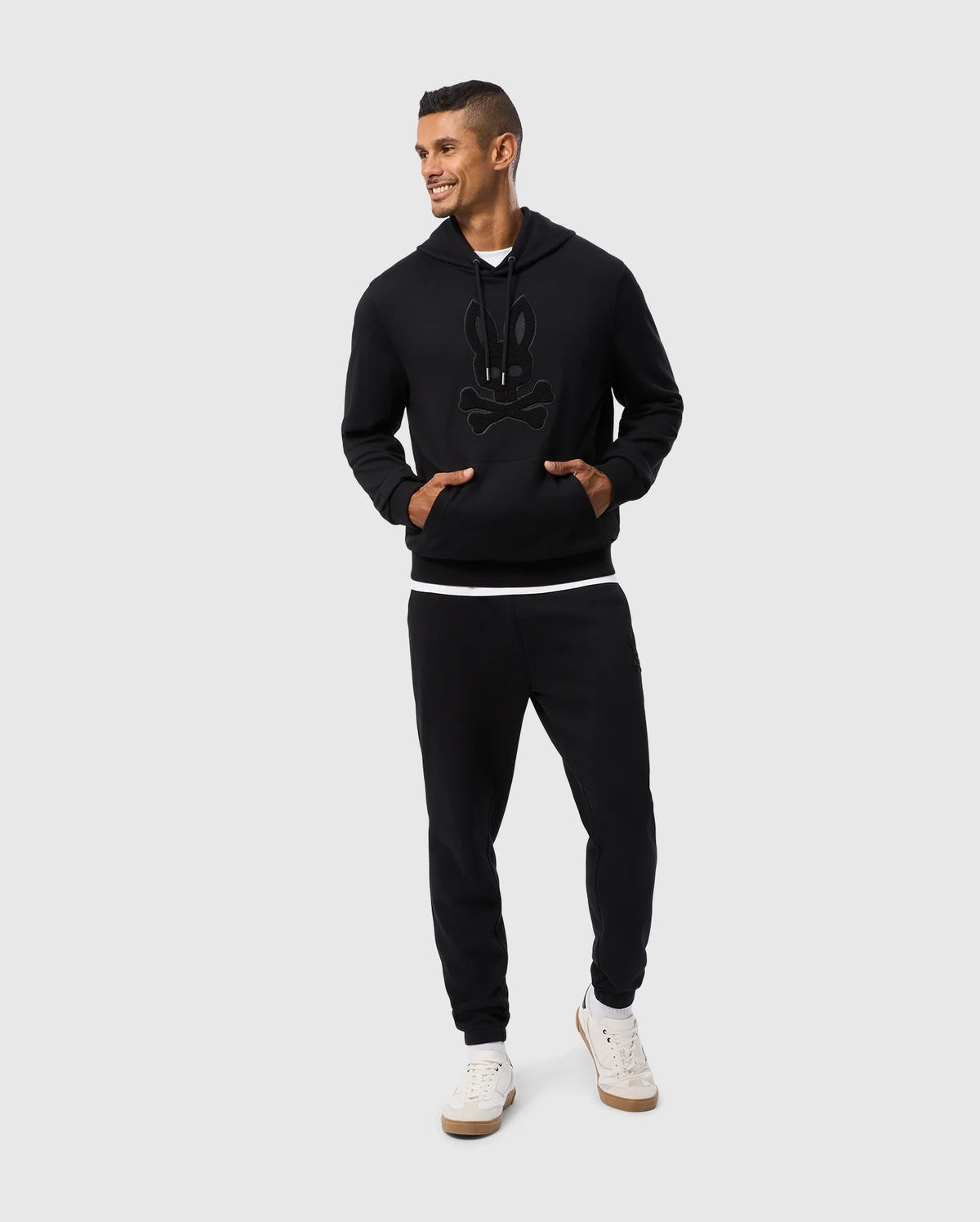 A man stands smiling, wearing a black hoodie featuring a high-pile chenille logo patch on the front, paired with Psycho Bunny's MENS PACIFIC CHENILLE JOGGER - B6P972D200. He has one hand in his pocket and is sporting white sneakers against a plain gray background.