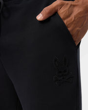 A close-up shows a person with a dark skin tone wearing Psycho Bunny's MENS PACIFIC CHENILLE JOGGER - B6P972D200, casually placing a hand in the pocket. This stylish bottom is adorned with an intricate embroidered design of a bunny skull with crossbones, set against a plain white background.