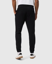A person wearing Psycho Bunny MENS PACIFIC CHENILLE JOGGERS - B6P972D200 in black and white sneakers stands with their back to the camera. These joggers are casual with an elastic waistband, a high-pile chenille logo patch, and a back pocket. The background is plain and light-colored.
