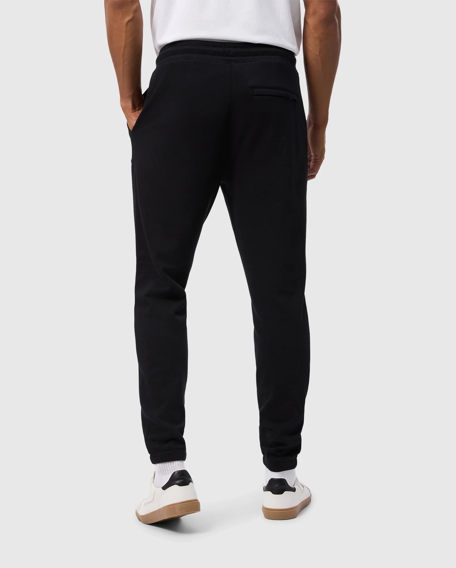 A person wearing Psycho Bunny MENS PACIFIC CHENILLE JOGGERS - B6P972D200 in black and white sneakers stands with their back to the camera. These joggers are casual with an elastic waistband, a high-pile chenille logo patch, and a back pocket. The background is plain and light-colored.