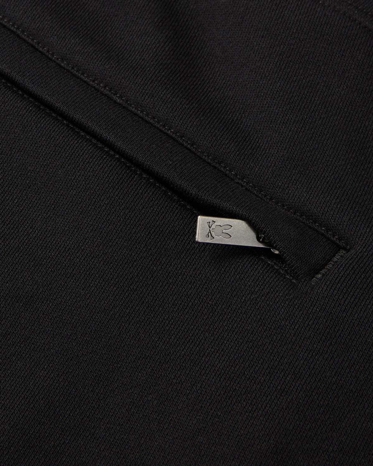 A close-up of black chenille fabric, featuring a metal zipper, evokes the style of a Psycho Bunny Pacific Chenille Jogger. The zipper pull is adorned with a subtle engraved logo design. The smooth, dark texture of this casual bottom contrasts elegantly with the slightly open zipper.