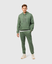 A person wearing a Psycho Bunny green hoodie adorned with a bunny graphic and a high-pile chenille logo patch pairs it with the MENS PACIFIC CHENILLE JOGGER - B6P972D200. They stand on a gray background, completing their casual look with off-white sneakers.