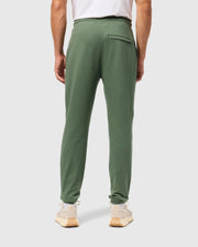 A person is wearing casual bottom Psyho Bunny Men's Pacific Chenille Joggers, paired with beige sneakers and a white t-shirt. They are standing with their back to the camera against a plain gray background.